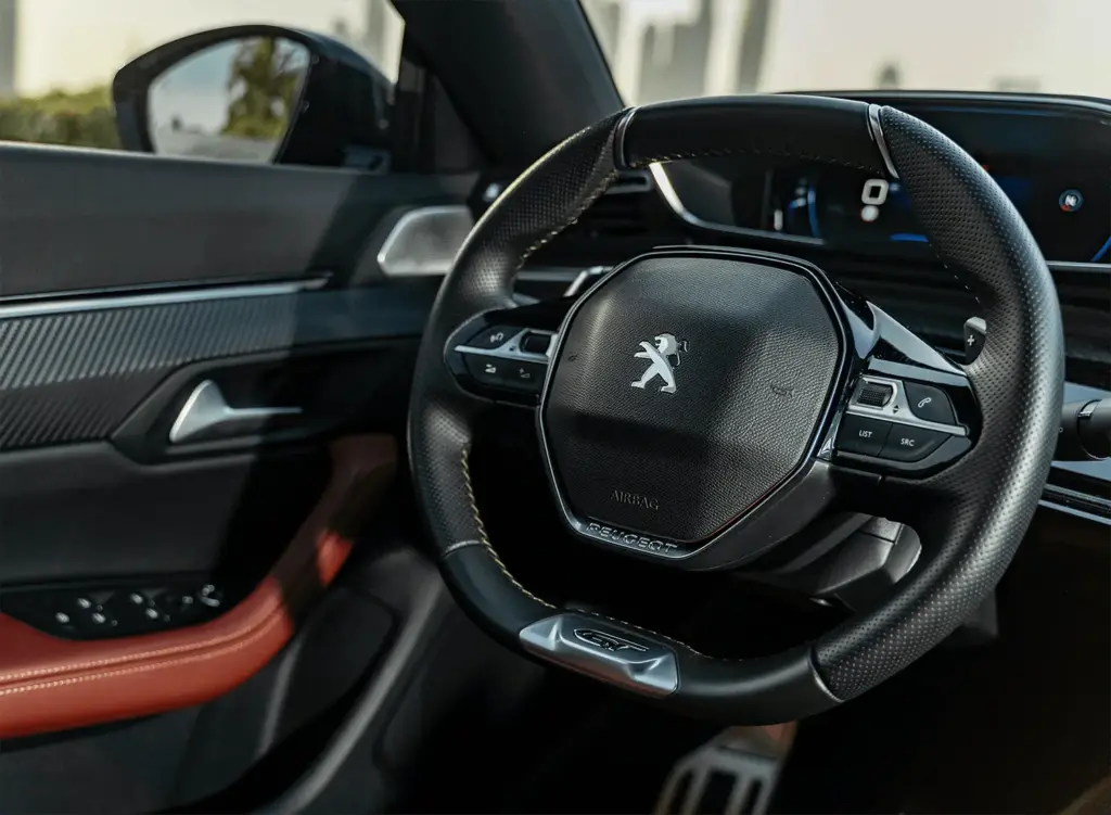 Rat i moderne bil viser Peugeot-logoet, omgivet af instrumentbræt med digitale målere og luksuriøs indretning. Læderbetrukket rat og kontrastfarver skaber elegant atmosfære. Sikrer komfortabel kørsel.