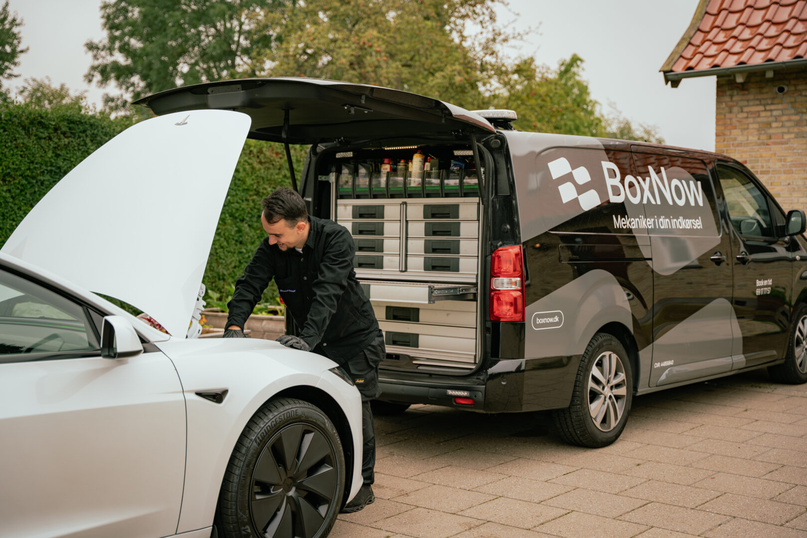 En mekaniker reparerer en bil med åben motorhjelm foran en sort varevogn. Tekst på varevognen: "BoxNow Mekaniker i din indkørsel, boxnow.dk." Haven og husets væg ses i baggrunden.