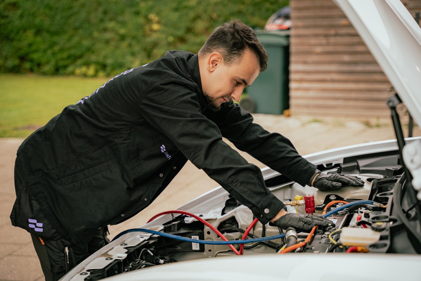 En mekaniker inspicerer en bilmotor under den åbne motorhjelm. Han bærer handsker og arbejder koncentreret. I baggrunden ses en græsplæne og en bygning.