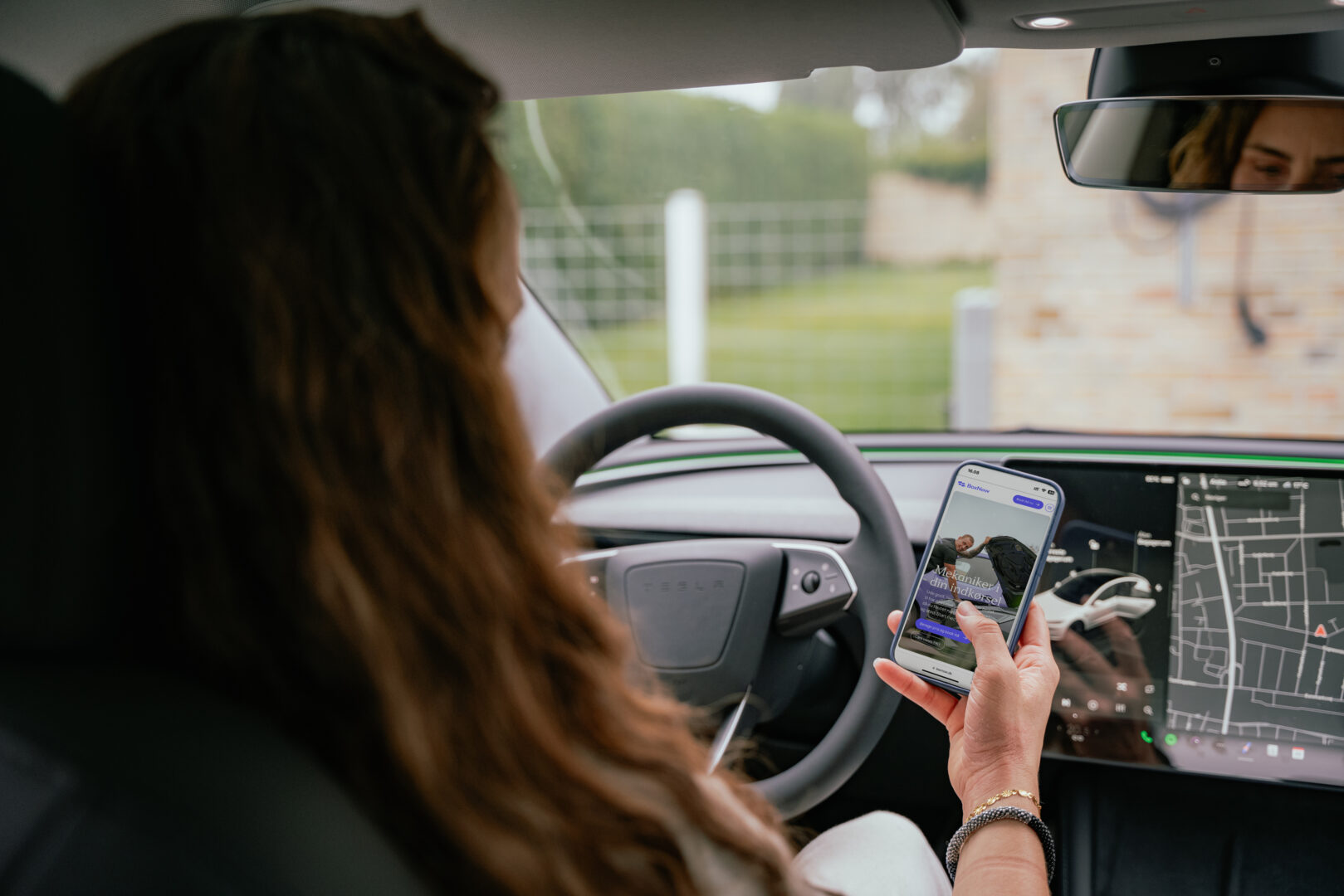 En person sidder i en bil og holder en smartphone, hvorpå der vises en mekaniker-app. Bilen har et moderne design med en detaljeret GPS-skærm i forruden.