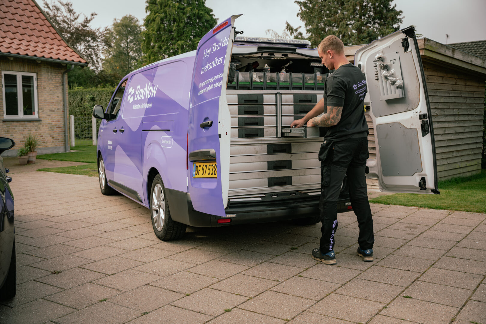 En mekaniker organiserer værktøjer i en åben varevogn fra "BoxNow" på en indkørsel. Bilen har reklame for mekanikerværksted. Omgivelserne er et boligområde med græs og træer. Tekst på bilen: "Hej! Skal du bruge mekaniker i indkørslen? Vi reparerer og servicerer biler, mens du får arbejdet gjort." "BoxNow mekaniker til indkørslen."