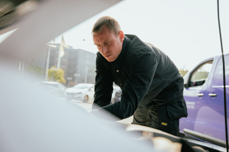 En mekaniker arbejder koncentreret på en bilmotor. Han bærer sort arbejdstøj. I baggrunden er der en blå varevogn og parkerede biler ved en vej i et bymiljø.