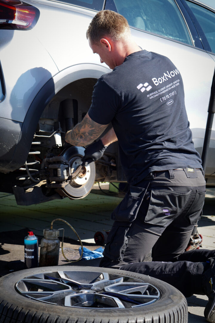BoxNow-mekaniker udfører bremsevæskeskift på elbil i indkørsel med specialværktøj, bremserens og opsamlingsflaske