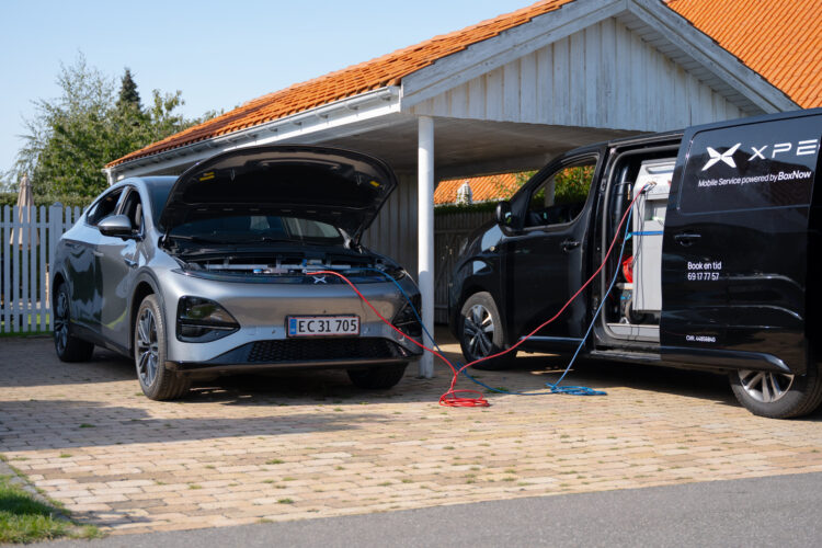 En grå bil med åben motorhjelm bliver forbundet til en sort varevogn via kabler i en indkørsel. Tekst på varevognen: "XPENG Mobile Service powered by BoxNow"