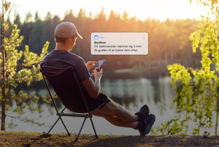 Manden sidder i en stol ved en sø, mens han bruger sin smartphone. En besked på skærmen siger: "Mail: BoxNow. Dit dækmønster nærmer sig 3 mm. Se guiden til at tjekke dem efter." Solen skinner gennem skovbaggrunden.